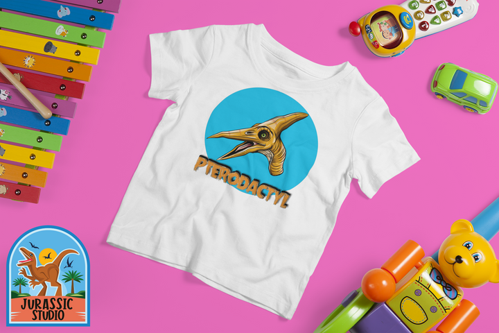 The Toddler Pterodactyl Head T-Shirt by Jurassic Studio features a playful dino design and “PTERODACTYL” text, displayed on a pink background with colorful toys—an ideal dinosaur fan gift! The Jurassic Studio logo is shown in the corner.