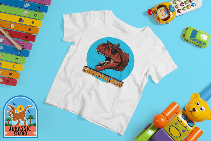 The Jurassic Studio Toddler Carnotaurus Head T-Shirt features a bold red dinosaur head and CARNOTAURUS text on blue, surrounded by colorful toys with the Jurassic Studio logo, making it a perfect gift for dino fans.