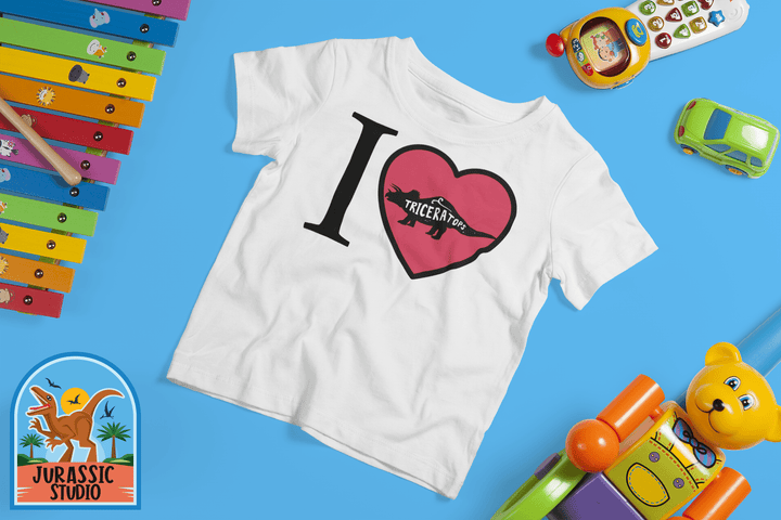 The Toddler I Love Triceratops T-Shirt by Jurassic Studio is displayed on a blue background with colorful toys, a xylophone, toy car, and plush bear nearby. The Jurassic Studio logo is in the bottom left corner.