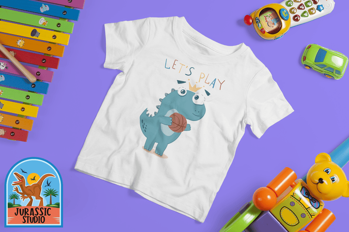 The Toddler Baby Dino Basketball T-Shirt by Jurassic Studio features a cute blue dinosaur holding a basketball with LETS PLAY text, surrounded by colorful toys on a purple background. The Jurassic Studio exclusive logo is at the bottom left.