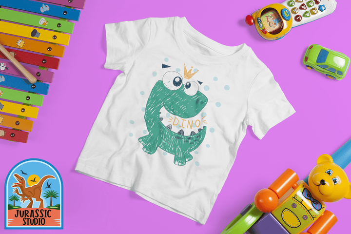 The Toddler Baby Green Dino T-Shirt by Jurassic Studio is a white 100% cotton tee featuring a crowned green cartoon dinosaur with toys on a purple background and the Jurassic Studio dino logo—an ideal gift for young dinosaur fans.