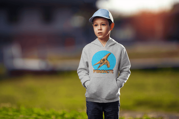 A young boy wears the Jurassic Studio Kids Pterodactyl Head Hoodie, featuring a bold pterodactyl graphic and PTERODACTYL text. He stands outdoors with his hands in his pockets, wearing a light blue cap against a blurred green background.