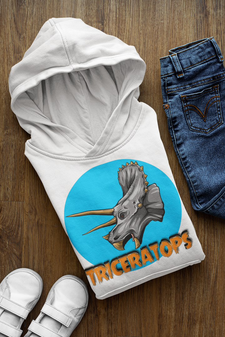 The Kids Triceratops Head Hoodie by Jurassic Studio, in white with a triceratops graphic and orange Triceratops text, is folded on a wooden floor beside blue jeans and white sneakers.