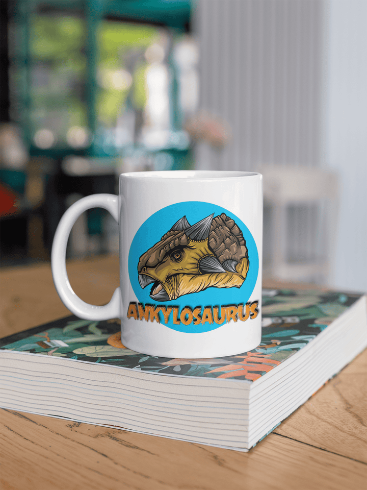 The Jurassic Studio Ankylosaurus Portrait Mug, with a vibrant ankylosaurus head and bold orange lettering, sits on a book on a wooden table in a bright, modern café.