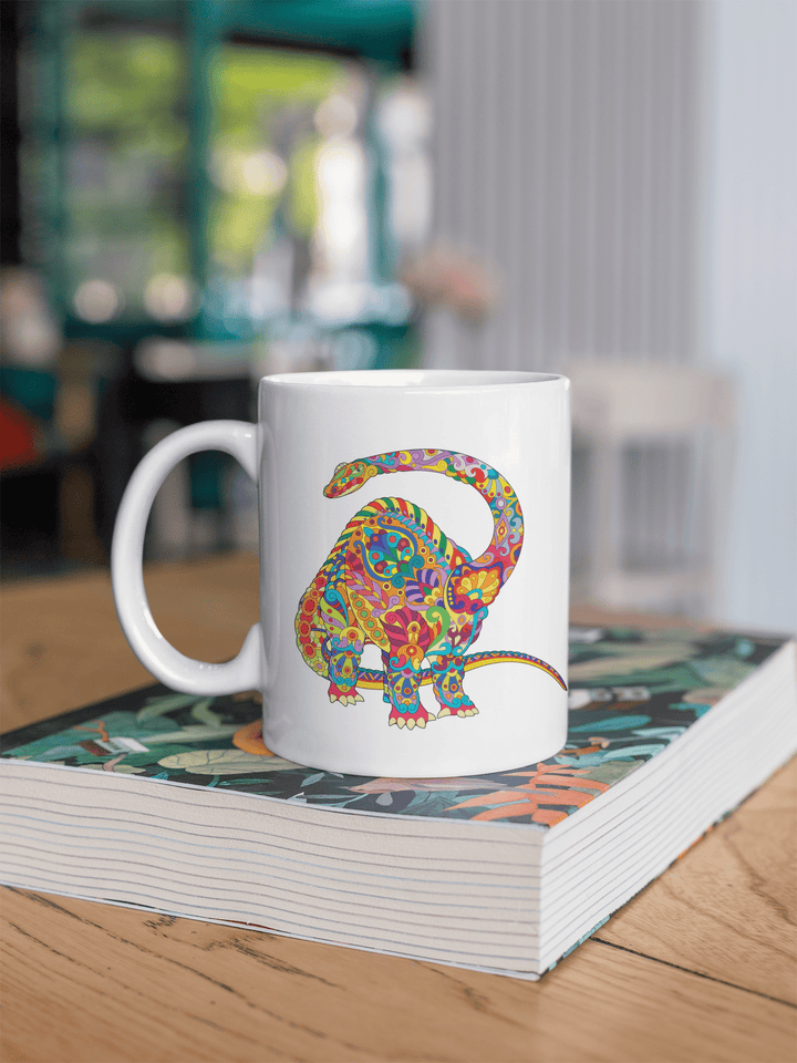 The Brontosaurus Zentangle Mug by Jurassic Studio, a white ceramic coffee mug with a vibrant patterned dinosaur design, rests on a leafy book atop a wooden table in a bright modern café.