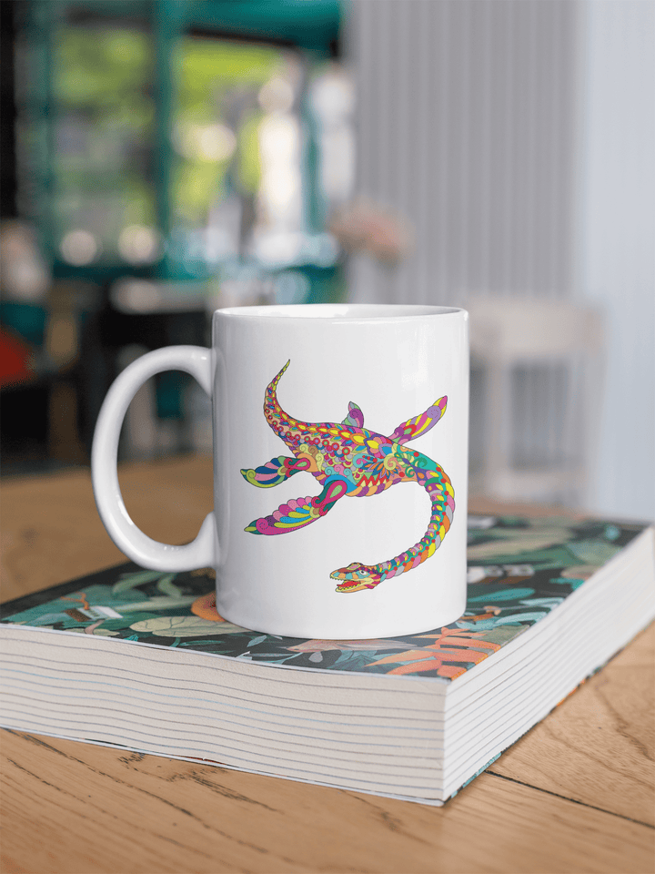 The Jurassic Studio Plesiosaurus Zentangle Mug, with its colorful prehistoric reptile design, rests on a thick patterned book atop a wooden table in a bright, modern café.