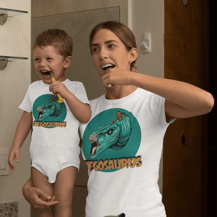 A woman and a boy in matching white Stegosaurus shirts brush their teeth by the mirror, smiling. Nearby hangs an adorable Jurassic Studio Toddler Stegosaurus Head Onesie, completing the dinosaur-themed scene.