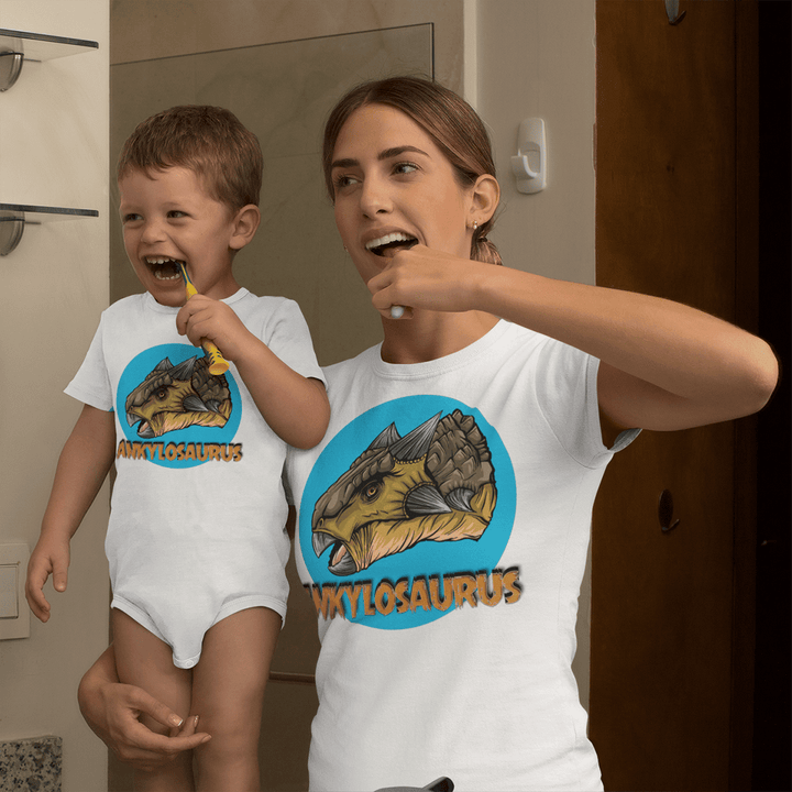 A woman and a young boy, both in Jurassic Studio apparel—the boy in a Toddler Ankylosaurus Head Onesie—brush their teeth together in front of a mirror, showing off their exclusive style.