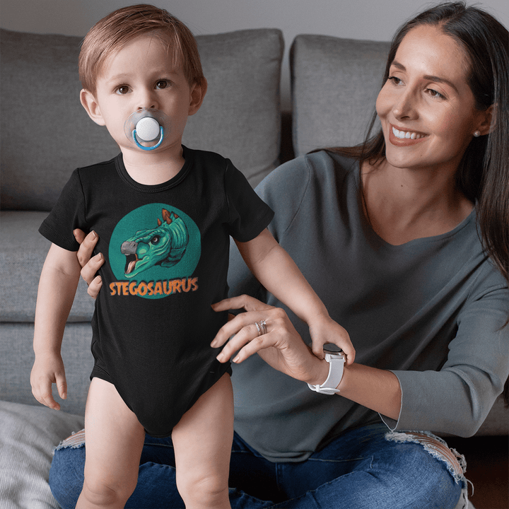 A smiling woman holds a toddler in a black Jurassic Studio Toddler Stegosaurus Head Onesie, which features a colorful cartoon stegosaurus and the word STEGOSAURUS. The child, with a pacifier, looks at the camera as they sit on an indoor couch.