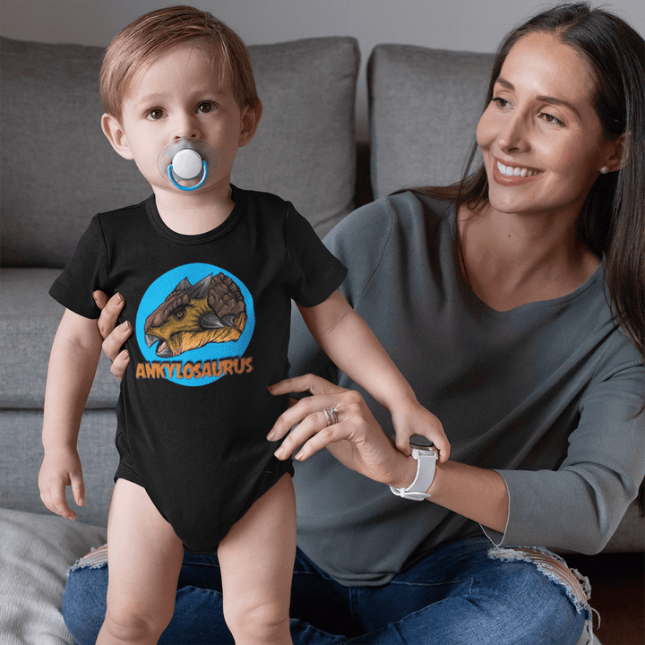 A smiling woman holds a toddler wearing the Jurassic Studio Toddler Ankylosaurus Head Onesie, featuring a colorful ankylosaurus design and ANKYLOSAURUS text. The pacifier-wearing child stands on a gray couch.