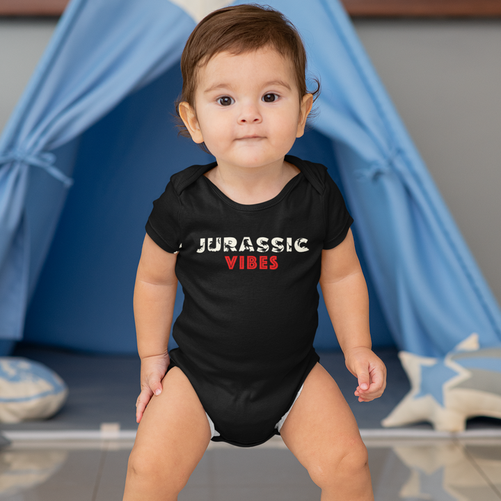 A toddler wears the Jurassic Studio Toddler Jurassic Vibes Onesie in front of a blue play tent, gazing at the camera with toys and cushions nearby—this cute dinosaur outfit adds playful charm to any young explorer’s day.