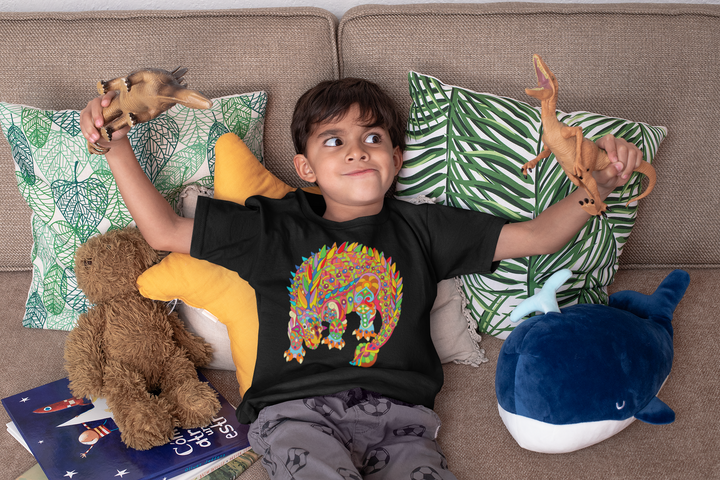 A young boy sits on a couch holding animal toys, surrounded by plushies and books. He wears the Kids Ankylosaurus Zentangle T-Shirt from Jurassic Studio—the ideal gift for dinosaur fans. Cushions have leafy and yellow patterns.