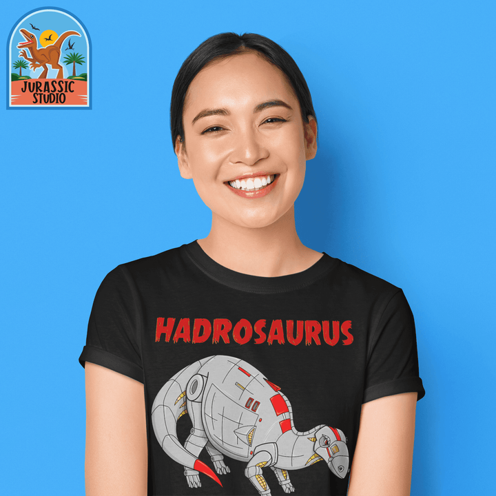 A woman smiles in a Women Robot Hadrosaur T-Shirt by Jurassic Studio, featuring a robot dinosaur design, standing against a blue background. The Jurassic Studio logo is displayed in the top left corner—a perfect gift for dinosaur fans.