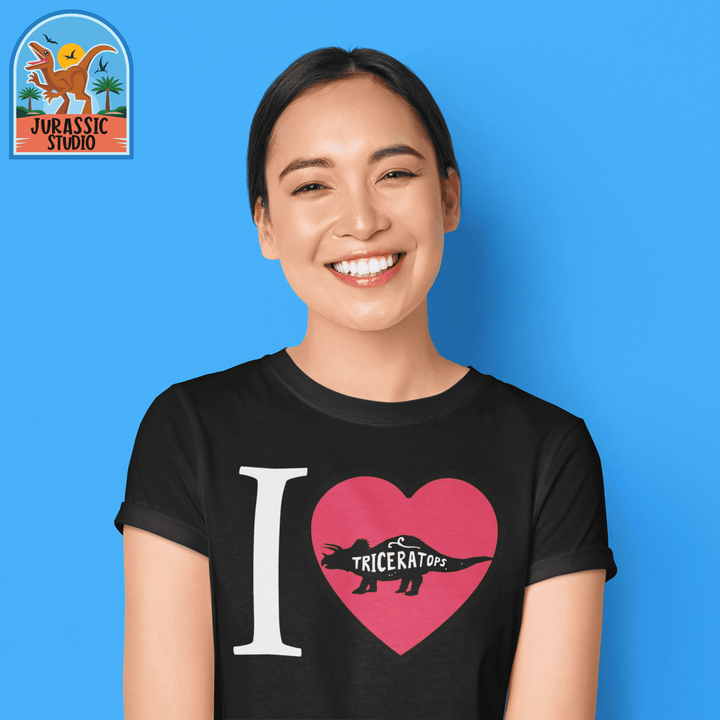 A smiling woman stands against a blue background wearing the Jurassic Studio Women I Love Triceratops T-Shirt, featuring a red heart and dinosaur silhouette with “I love Triceratops”—a perfect dinosaur gift.