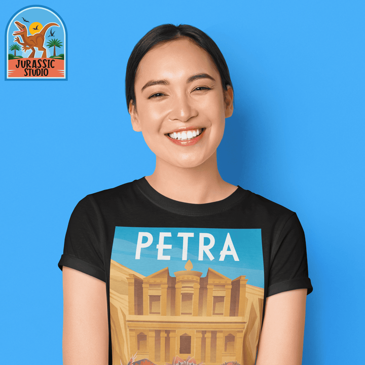 A smiling woman in a slim fit Women Petra T-Shirt by Jurassic Studio poses against a blue background. The shirt showcases Petra’s Treasury, and the Jurassic Studio dinosaur logo is visible in the top left corner.