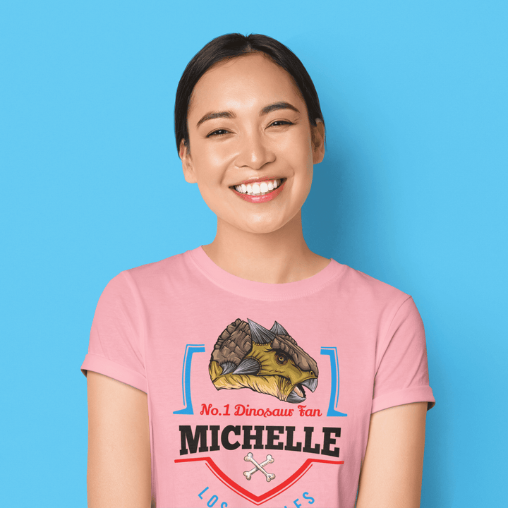 Smiling woman with dark hair in a ponytail wears the Jurassic Studio N.1 Ankylosaurus Fan Custom Women T-Shirt, personalized with “No.1 Dinosaur Fan Michelle” and a dinosaur illustration, set against a blue background.
