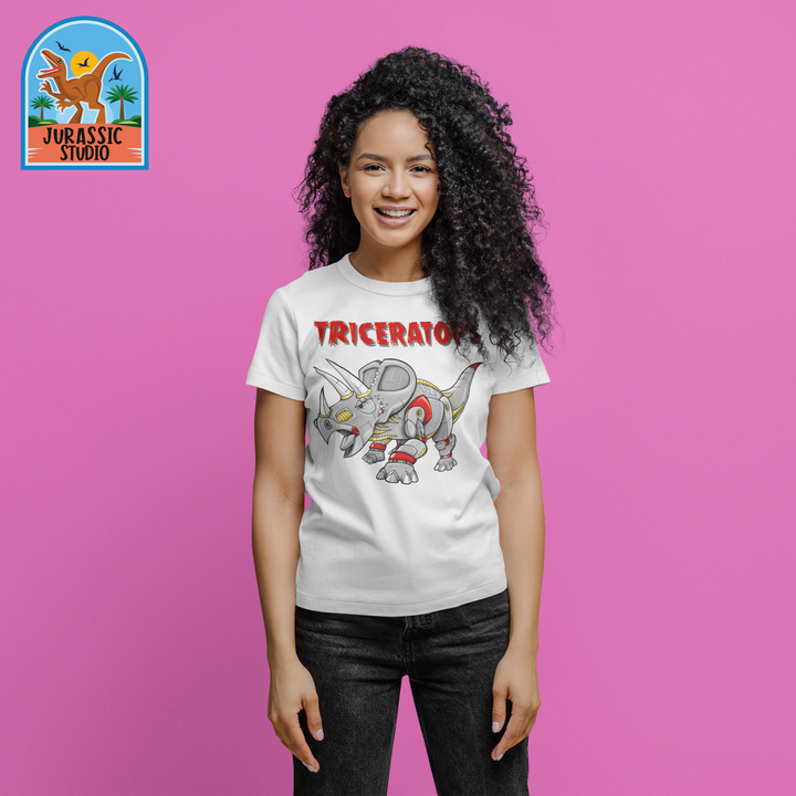 A smiling person with curly hair stands against a pink background, wearing the Jurassic Studio Women Robot Triceratops T-Shirt featuring a robot triceratops and TRICERATOR, with the Jurassic Studio logo in the top left corner.