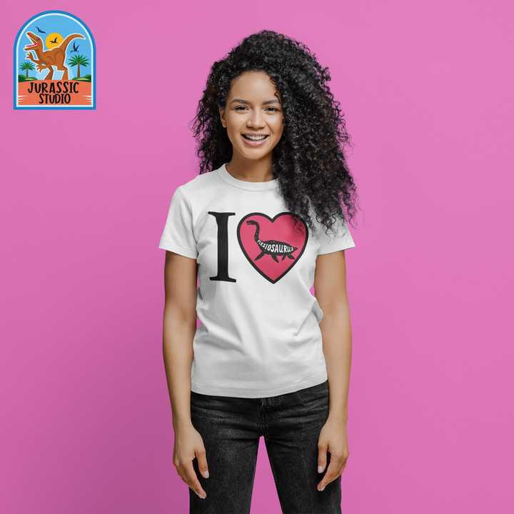 A smiling woman poses against a pink background wearing the Jurassic Studio Women I Love Plesiosaurus T-Shirt, perfect for dinosaur fans. The top left displays the Jurassic Studio logo with a dinosaur and palm trees.