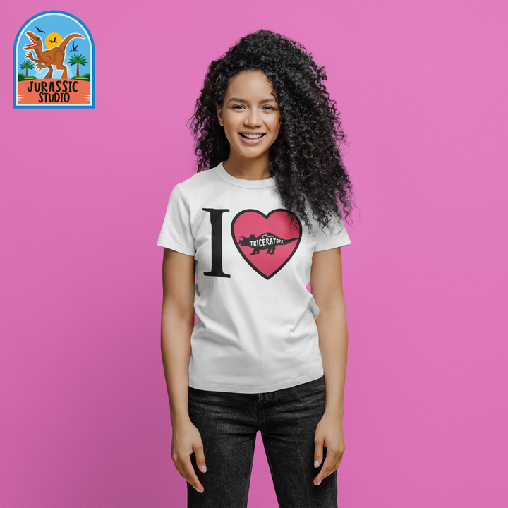 A smiling woman with curly hair stands before a pink background, wearing the Women I Love Triceratops T-Shirt by Jurassic Studio. The brands dinosaur and palm trees logo appears in the top left corner.