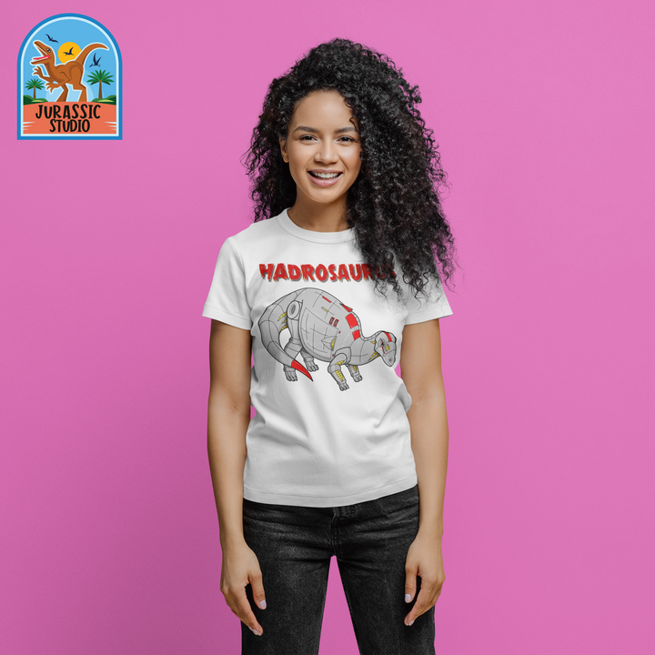 A smiling woman with curly hair wears the Jurassic Studio Women Robot Hadrosaur T-Shirt, featuring a cartoon hadrosaur and HADROSAURUS, against a pink background with the Jurassic Studio logo in the top left.