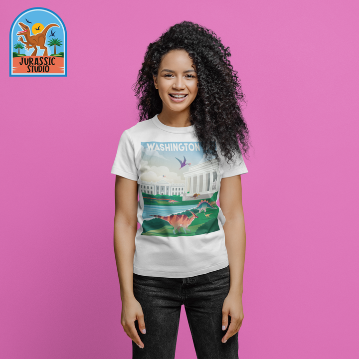 A smiling woman stands against a pink background, wearing the Jurassic Studio Women Washington DC T-Shirt with a dinosaur and White House graphic—an ideal gift for dinosaur fans. The exclusive Jurassic Studio logo is shown in the top left corner.