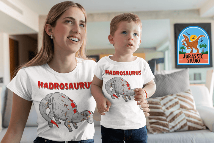 A smiling woman and young boy sit indoors, both in matching Jurassic Studio Women Robot Hadrosaur T-Shirts featuring a cartoon hadrosaurus—an ideal dinosaur fan gift. The exclusive Jurassic Studio logo appears in the background.