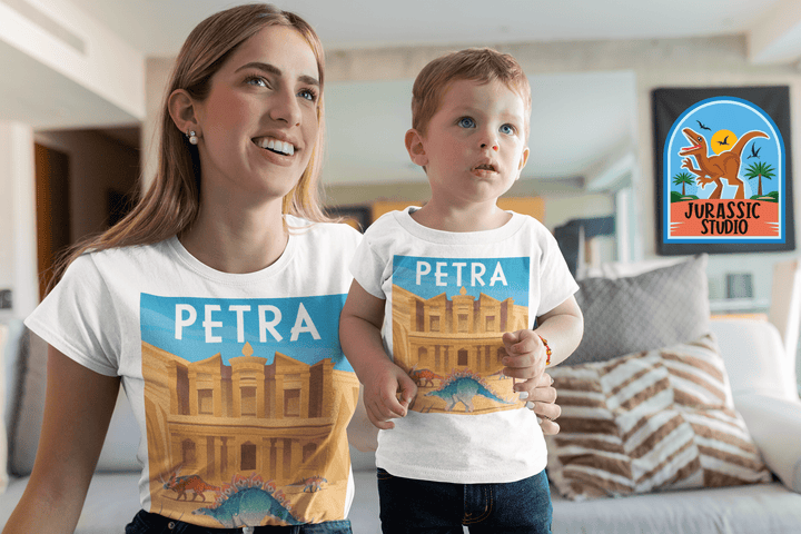 A woman and a young boy sit on a couch indoors wearing Jurassic Studio’s Women Petra T-Shirts, each featuring Petra with dinosaurs. A cushion in the background displays the Jurassic Studio exclusive logo.