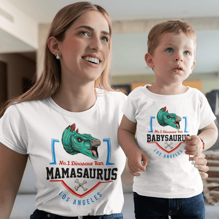 A smiling woman and child stand indoors, wearing matching Jurassic Studio N.1 Stegosaurus Fan Custom Women T-Shirts with cartoon dinosaur heads—hers says MAMASAURUS, the childs says BABYSAURUS. Both look happy together.
