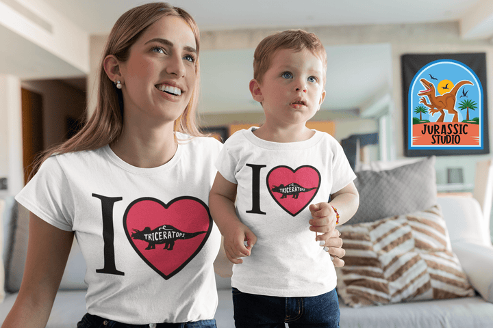 A smiling woman and child wear matching Jurassic Studio Women I Love Triceratops T-Shirts with dinosaur designs, sitting indoors on a couch with the colorful brand logo behind them—a perfect dinosaur-themed gift moment.