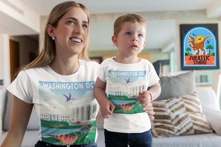 A smiling woman and young boy wear matching Jurassic Studio Women Washington DC T-Shirts with a dinosaur and monuments design, sitting on a couch—an exclusive, perfect gift for any dinosaur fan!.