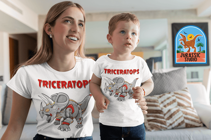 A woman and child sit together on a couch, both wearing matching white Jurassic Studio Women Robot Triceratops T-Shirts featuring a cartoon triceratops. A colorful Jurassic Studio sign is visible on the wall behind them.