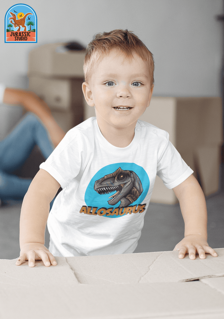 A smiling young child wears a white Toddler Allosaurus Head T-Shirt by Jurassic Studio and leans on a cardboard box indoors, with more boxes and a seated adult in the background—a perfect gift for young dinosaur fans.