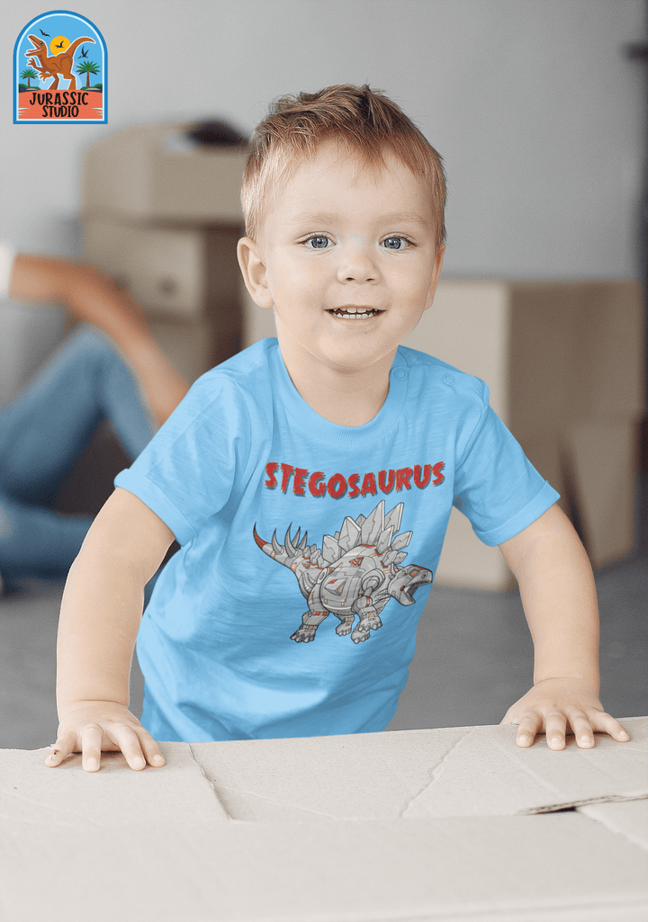A smiling child wearing the Toddler Robot Stegosaurus T-Shirt by Jurassic Studio plays indoors near cardboard boxes, with an adult in the background. Ideal gift for dinosaur fans! Jurassic Studio logo is visible in the top left corner.