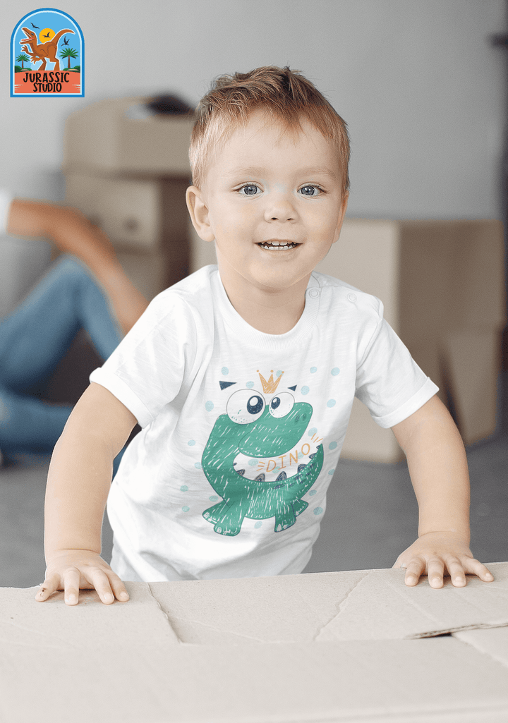 A smiling toddler wears the Jurassic Studio Toddler Baby Green Dino T-Shirt, made of 100% cotton and decorated with a cute cartoon dinosaur and DINO—a perfect gift for young dino fans. The Jurassic Studio logo appears in the top left corner.