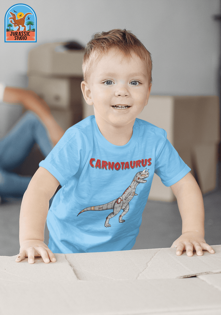 A smiling young boy stands indoors near cardboard boxes, wearing a light blue Jurassic Studio Toddler Robot Carnotaurus T-Shirt with a cartoon dinosaur—a perfect gift for any dino fan. A blurred adult is in the background.