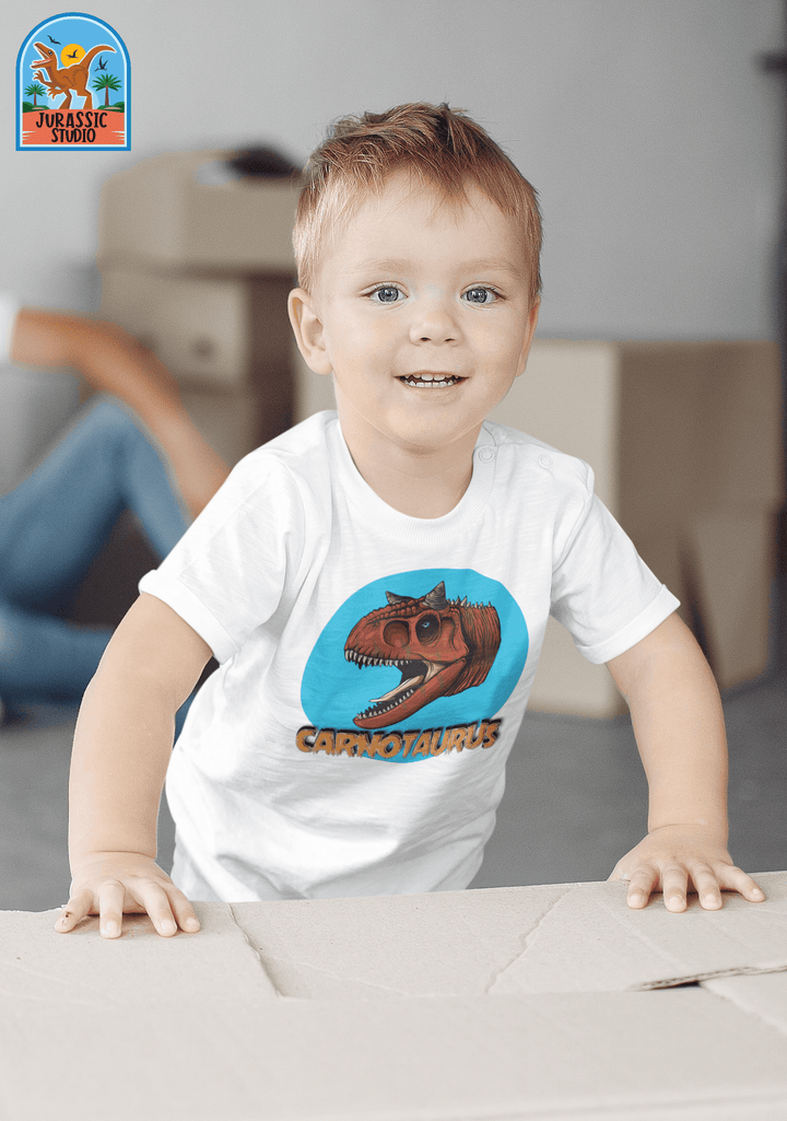 A young child with light brown hair smiles while wearing a Jurassic Studio Toddler Carnotaurus Head T-Shirt. Cardboard boxes and a seated adult are in the background, with the Jurassic Studio logo in the top left corner.