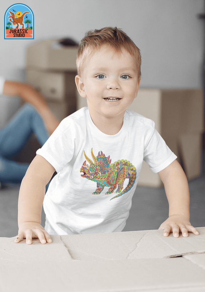 A smiling toddler with short light brown hair wears the Jurassic Studio Toddler Triceratops Zentangle T-Shirt—a great dinosaur fan gift—while leaning on a cardboard box, with another person and more boxes in the background.
