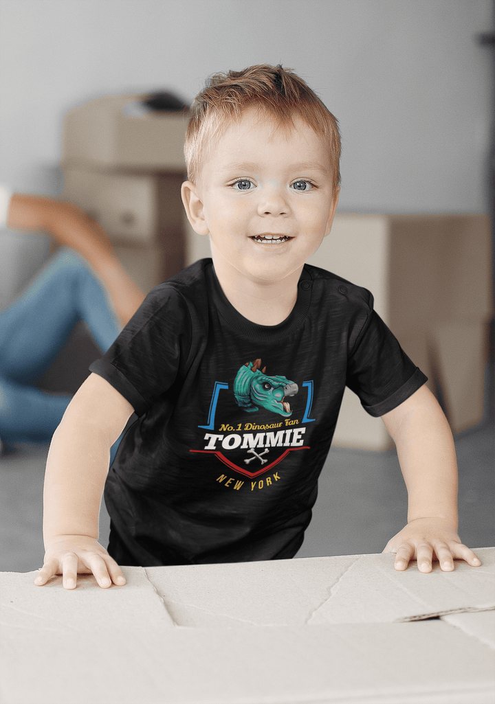 A smiling young boy with light hair wears a Jurassic Studio N.1 Stegosaurus Fan Custom Toddler T-Shirt, featuring a green dinosaur and custom name. He leans on a cardboard box, with blurred boxes in the background.