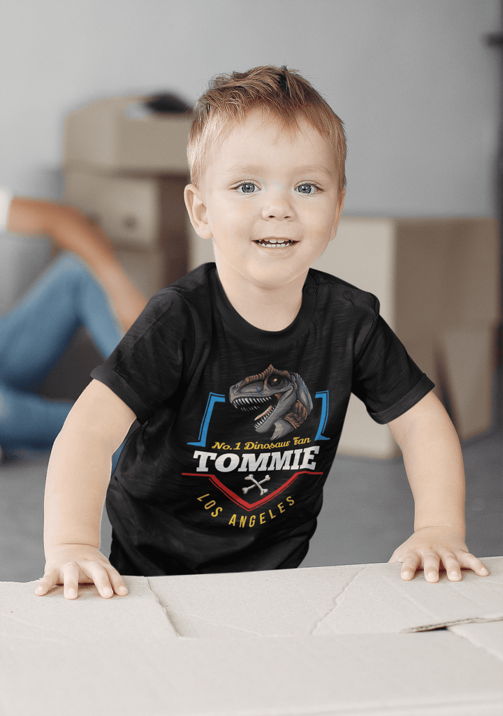 A smiling young boy with light brown hair wears a Jurassic Studio N.1 Allosaurus Fan Custom Toddler T-Shirt personalized with Tommie. He stands indoors, leaning on a cardboard box, with blurred boxes in the background.