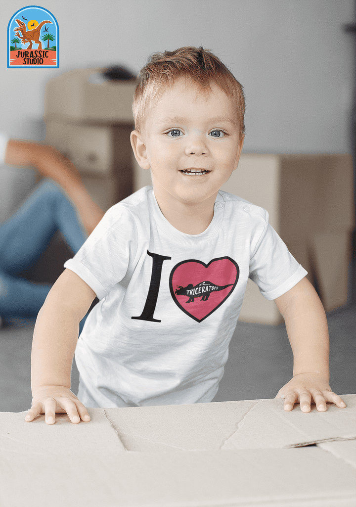 A happy child in a white Toddler I Love Triceratops T-Shirt by Jurassic Studio stands near a cardboard box, with moving boxes softly blurred in the background and the Jurassic Studio logo in the top left corner.