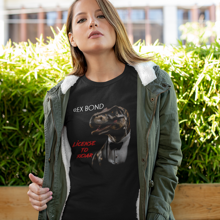 Outdoors, a woman wears the Jurassic Studio Women Rex Bond T-Shirt featuring a dinosaur in a tuxedo with Rex Bond and License to Roar in red—an ideal gift for dinosaur fans. Green foliage and a building are visible in the background.