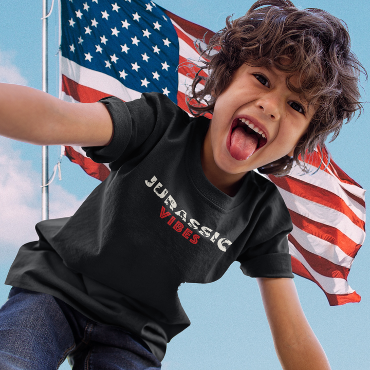 A cheerful boy with messy hair sticks out his tongue and smiles, wearing the Jurassic Studio Kids Jurassic Vibes T-Shirt—perfect for dinosaur fans. An American flag waves in the background against a blue sky.